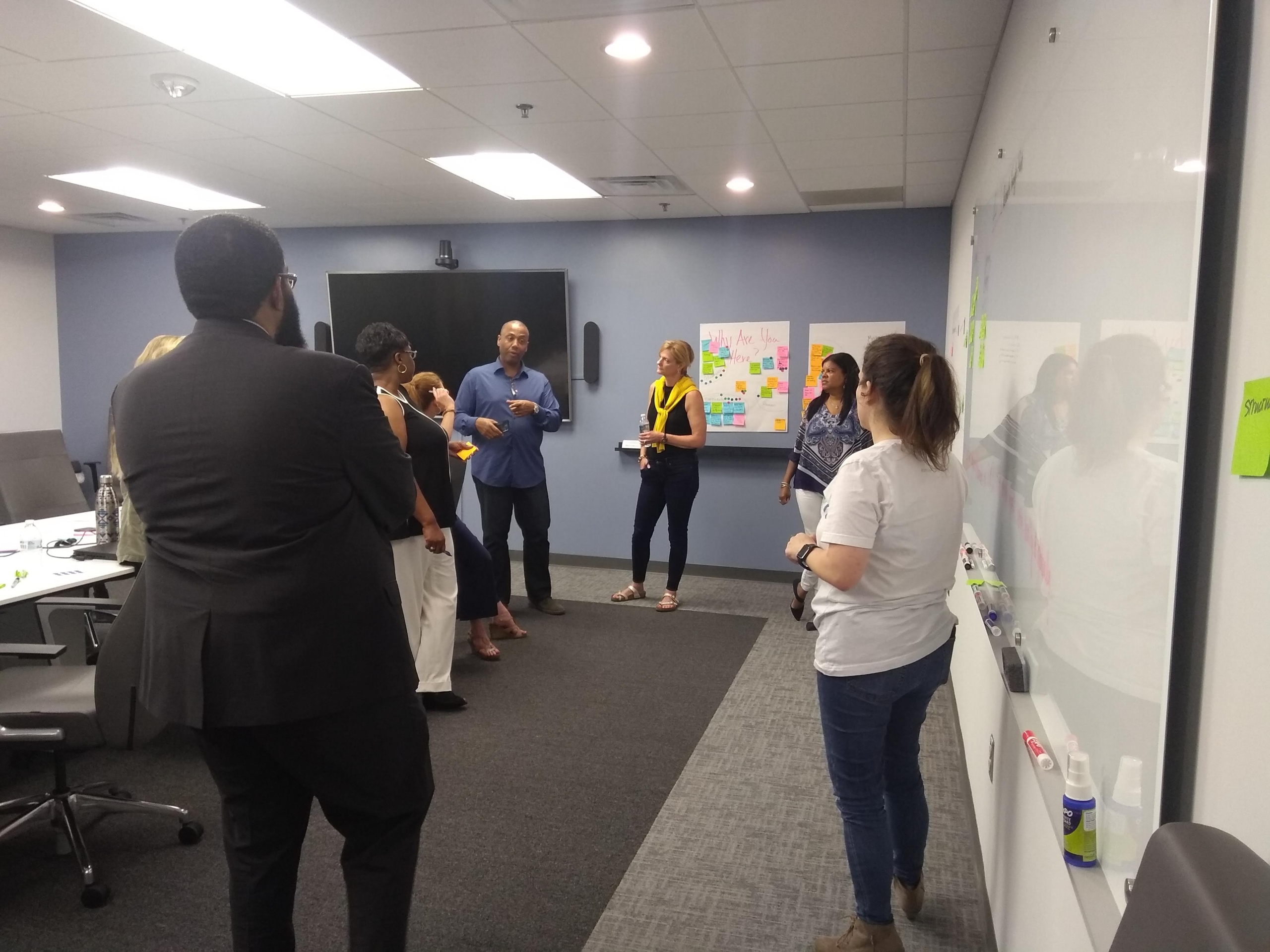 Seven people standing in a conference room talking in front of a whiteboard with sticky notes.